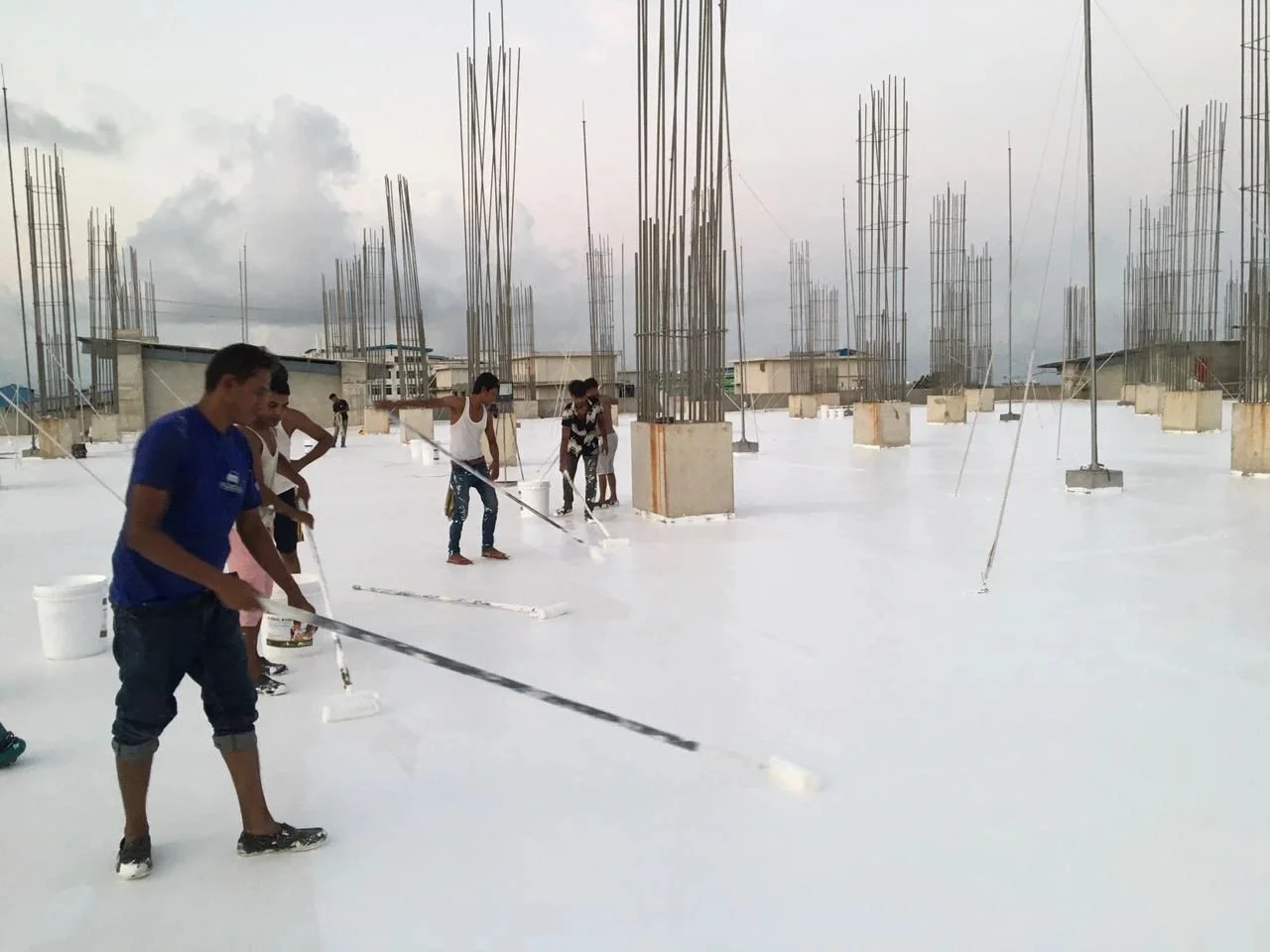 Workers applying white acrylic waterproofing coating with rollers on a roof slab